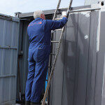 A painter painting a large container while working from a set of ladders